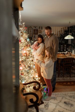 Family of four admiring a decorated Christmas tree with glowing lights in a cozy, warmly lit living room.