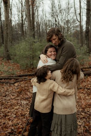 Family of four sharing a warm group hug in an autumn forest with fallen leaves and bare trees.