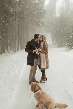 Couple kissing in snowy forest holding ultrasound photos with golden dog nearby, winter pregnancy announcement.