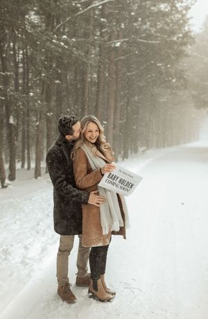 Couple embracing in snowy forest holding Baby Holden pregnancy announcement sign during winter snowfall.