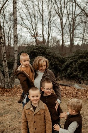 A woman poses outdoors in autumn with four young boys wearing coordinating brown jackets among bare trees.