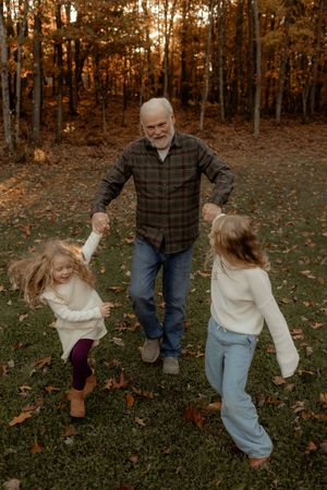 Grandfather holding hands with two young girls dancing outdoors among autumn leaves and trees.