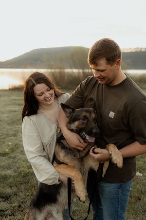 A smiling couple holds their German Shepherd dog outdoors near a lake at sunset.