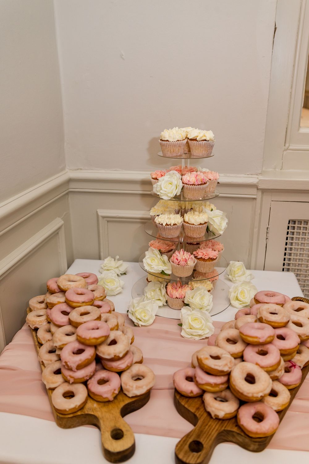 Pink glazed donuts on wooden boards beside a tiered cupcake stand with roses at a wedding reception.