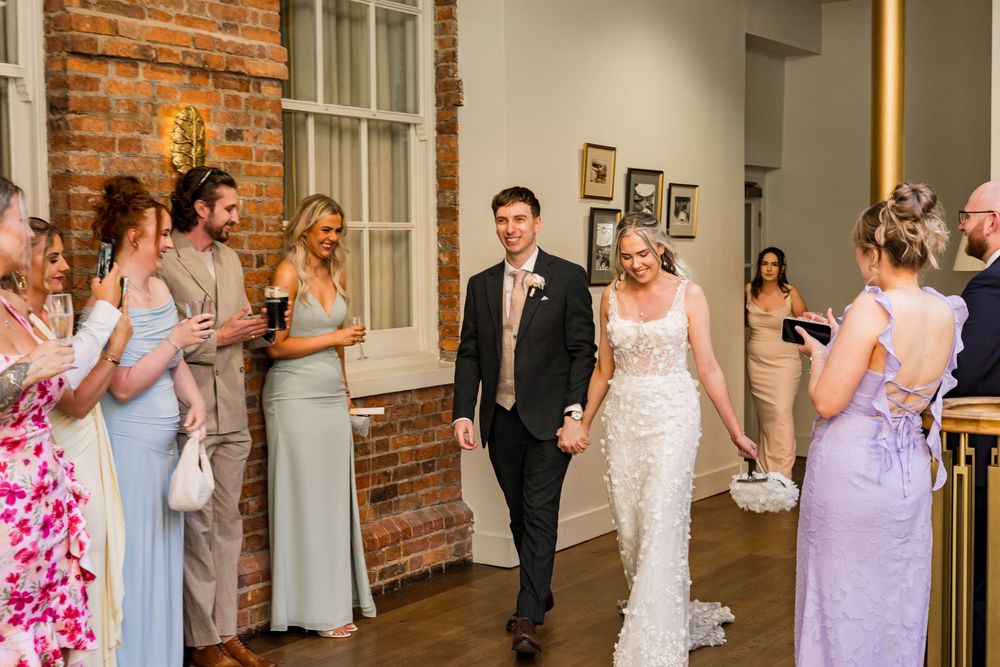 Bride and groom walk hand in hand past smiling guests in an elegant brick-walled wedding venue.