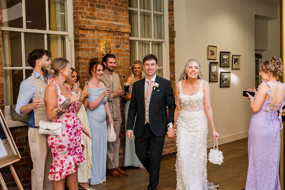 Bride and groom make grand entrance at wedding reception while guests cheer and applaud in stylish venue.
