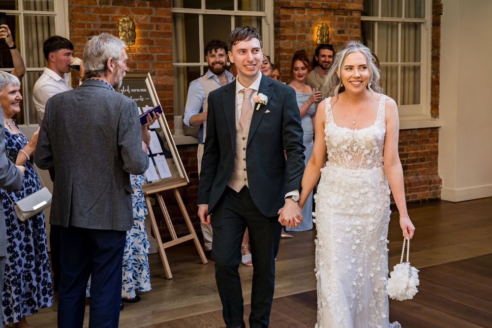 Smiling bride in floral wedding gown and groom in navy suit walk hand-in-hand past applauding wedding guests.
