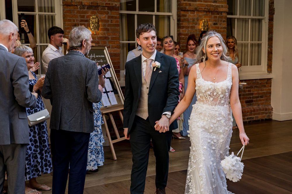 Newlywed couple walking hand in hand at wedding reception in elegant venue with guests.