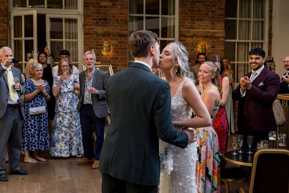 Bride and groom share their first dance kiss surrounded by applauding wedding guests.