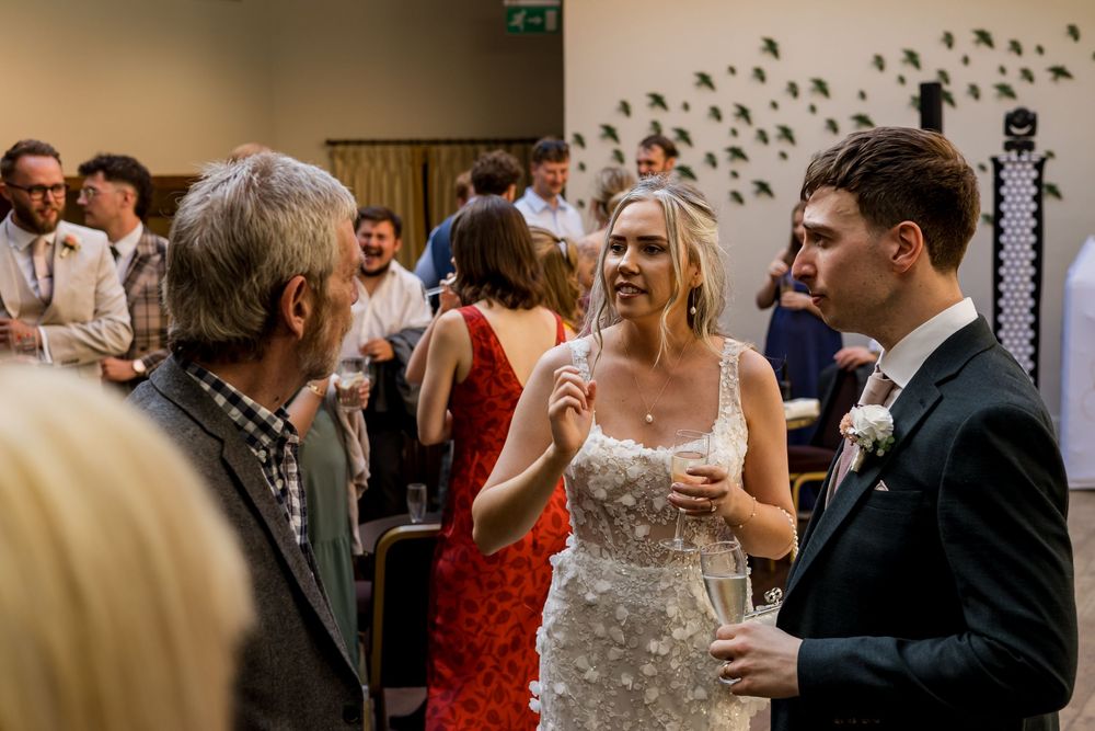Bride in floral gown mingles with wedding guests during reception cocktail hour in modern venue.