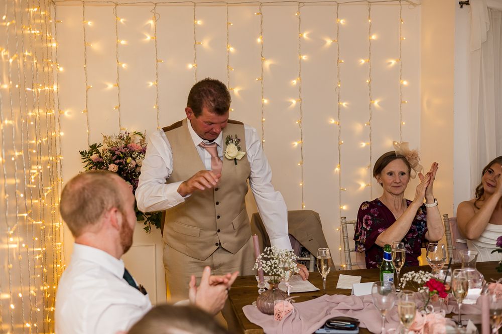 Groom giving a wedding speech at a decorated reception venue with fairy lights and guests applauding.
