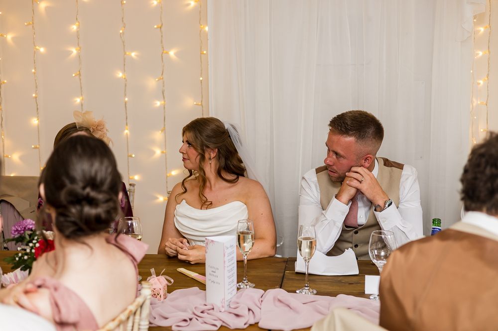 Bride and groom laughing at wedding reception table with fairy lights backdrop and pink table decorations.