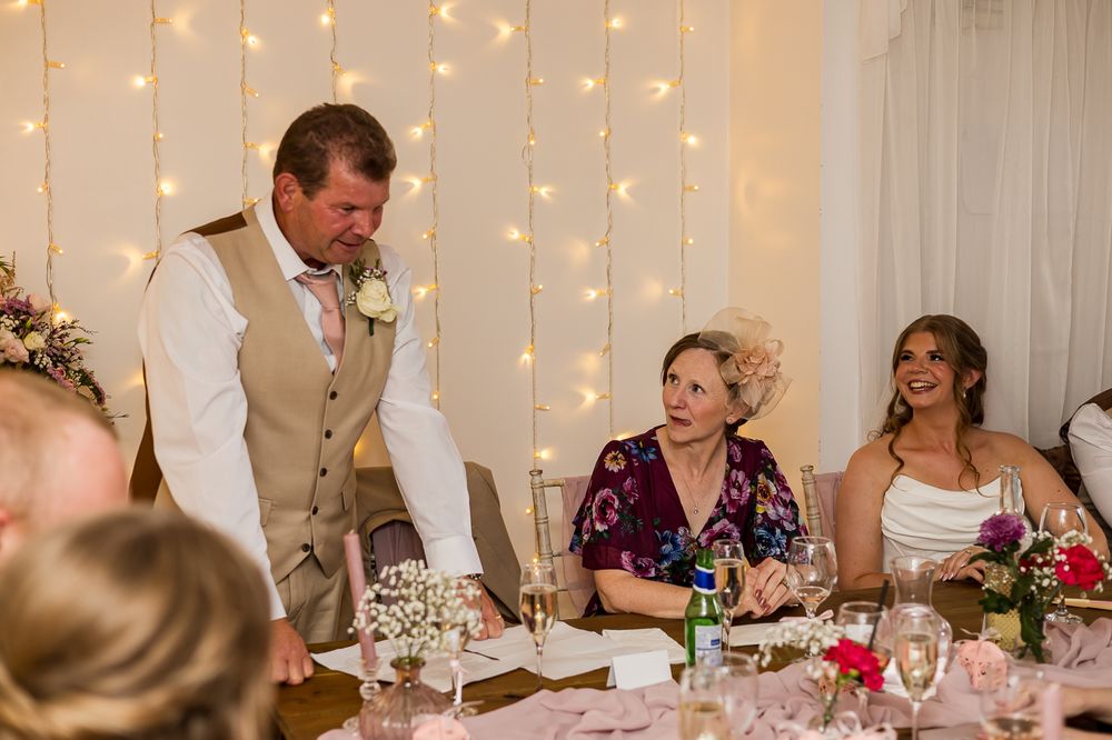 A groom in a tan vest gives a wedding speech as the bride in white and a guest in purple smile at a decorated reception table.