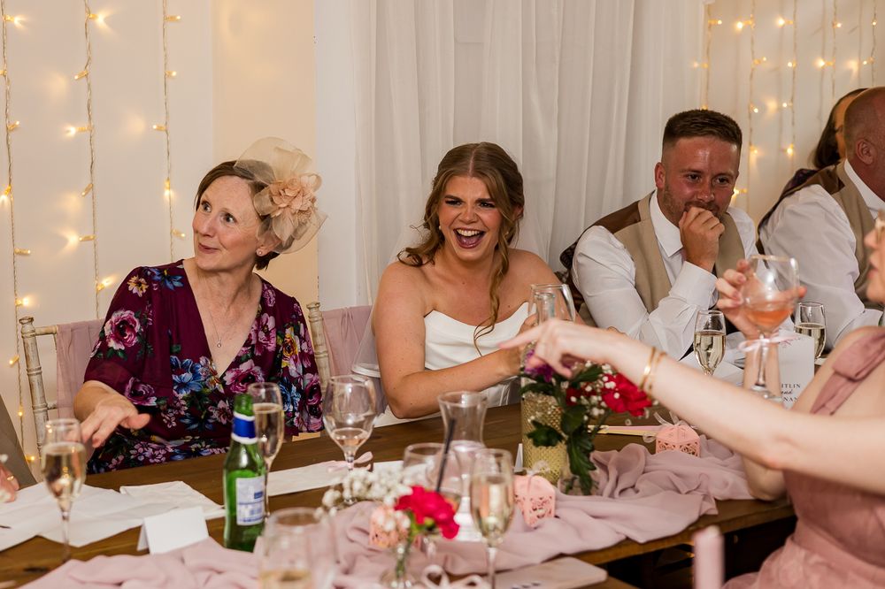 Bride laughing joyfully at wedding reception table with guests, fairy lights and floral decorations in background.