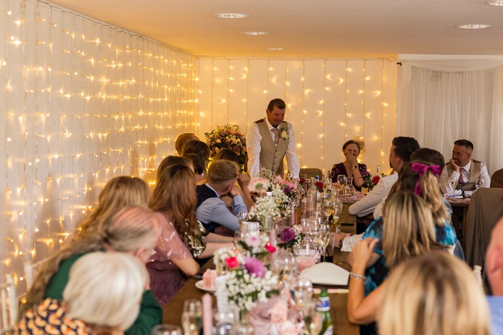 Wedding reception dinner with guests seated at floral tables, fairy light backdrop, and groom giving a speech.