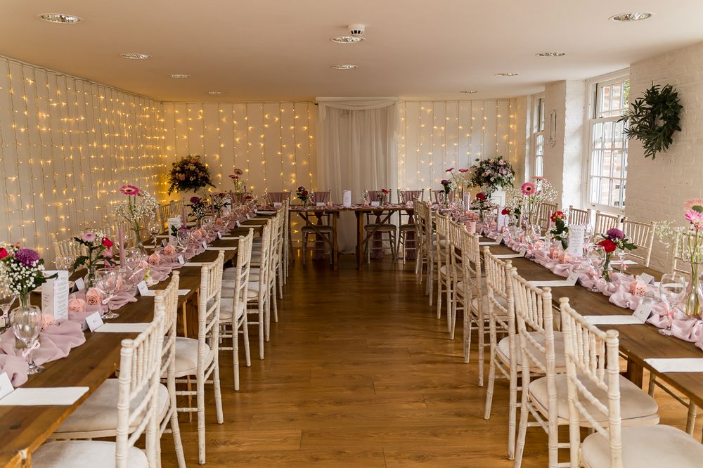 Elegant wedding reception room with fairy light backdrop, white Chiavari chairs, and pink floral centerpieces on rustic wooden tables.