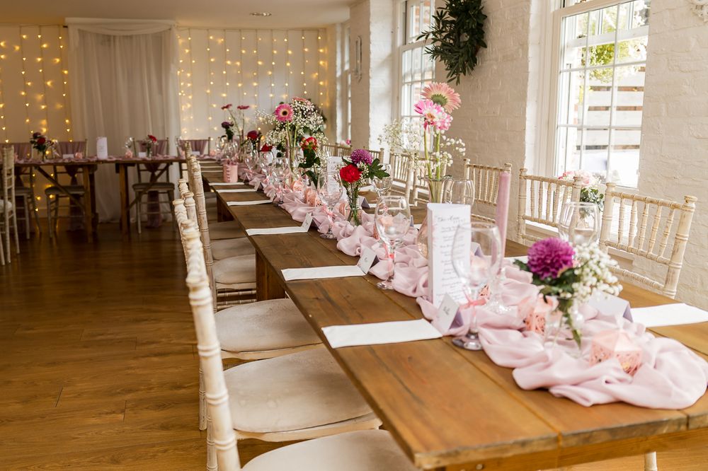 Elegant wedding reception with long wooden tables, pink floral arrangements, chiavari chairs, and fairy lights in a bright venue.