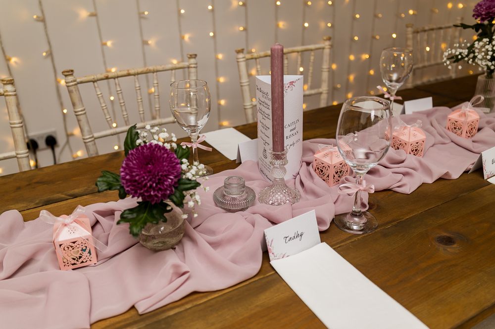 Elegant wedding table setting with pink runner, purple flowers, candles, and fairy lights backdrop.