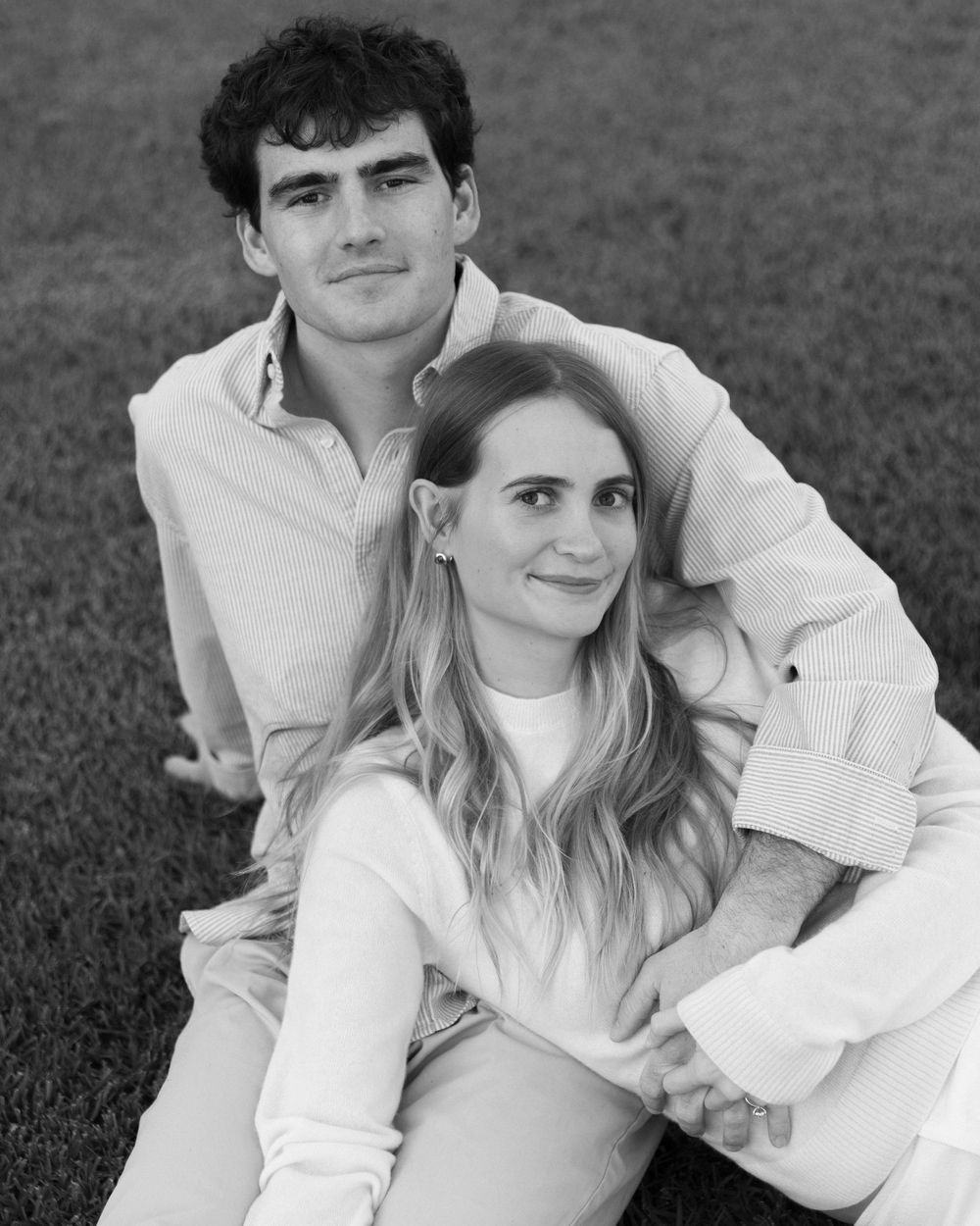 Black and white photo of a young couple sitting on grass, smiling warmly in casual attire.
