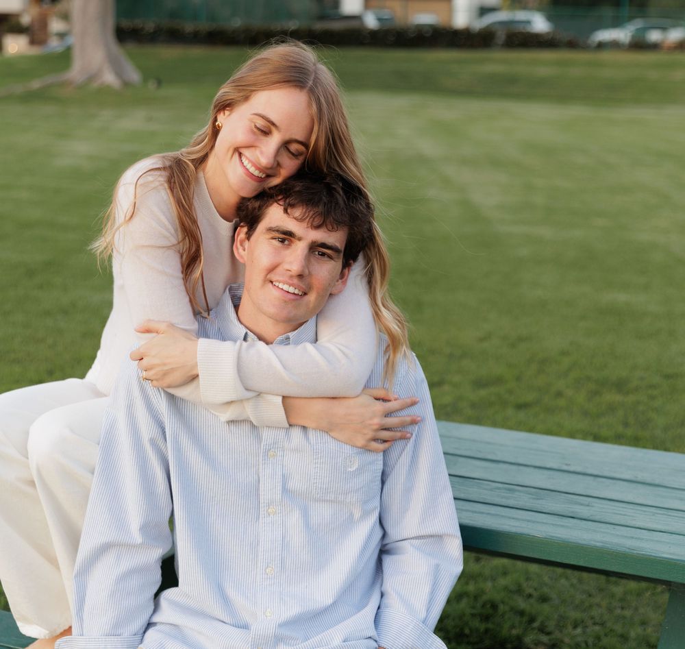 A young couple poses on a green bench outdoors, woman hugging man from behind, both smiling happily.