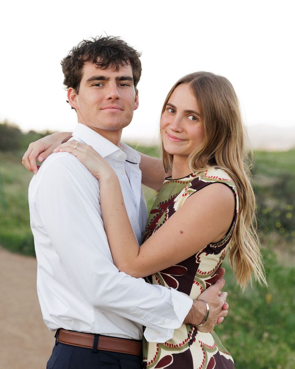 A young couple embracing outdoors, smiling warmly in casual attire against a scenic coastal backdrop.