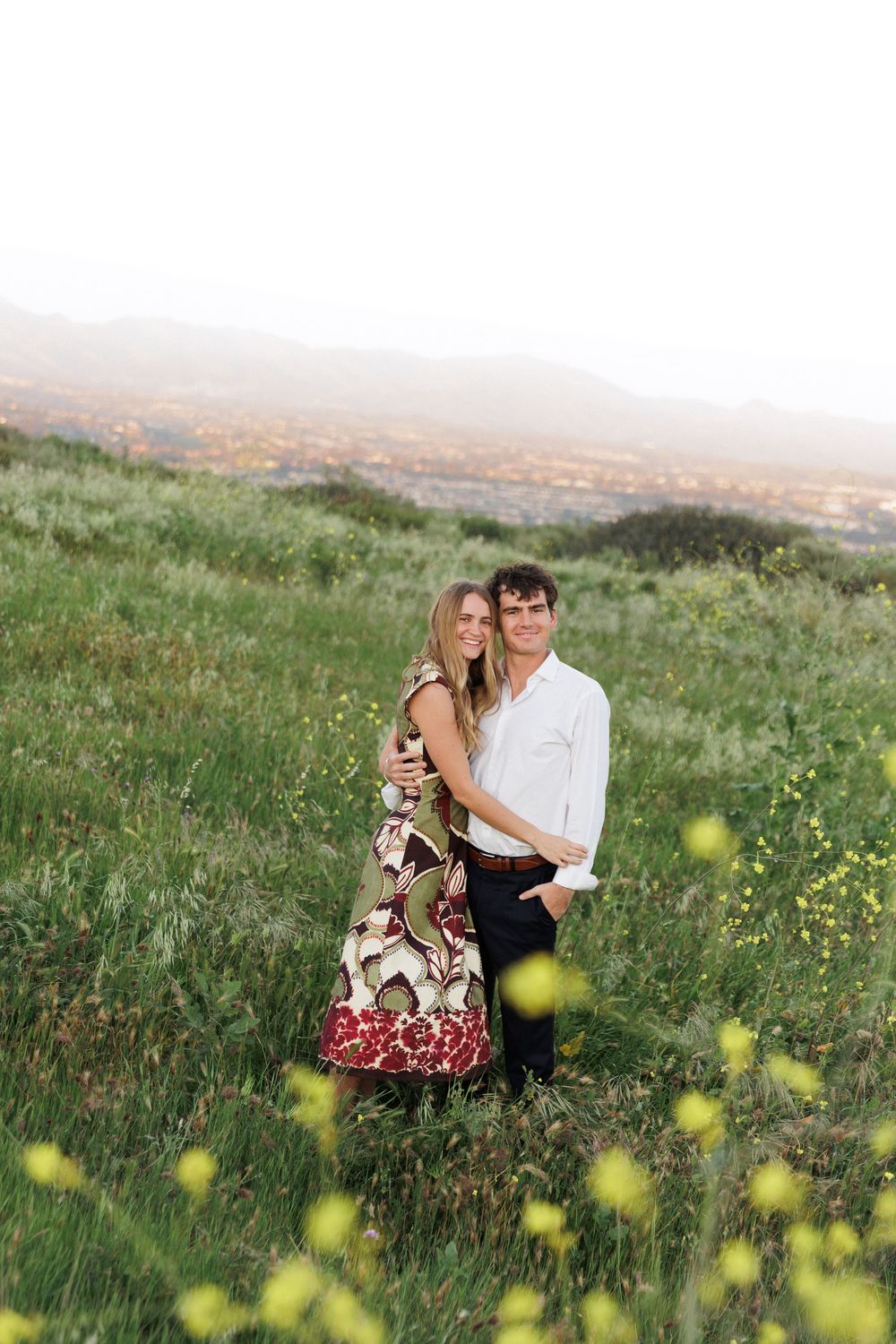 Couple hugging in a lush green meadow with yellow wildflowers and a scenic valley view behind them.