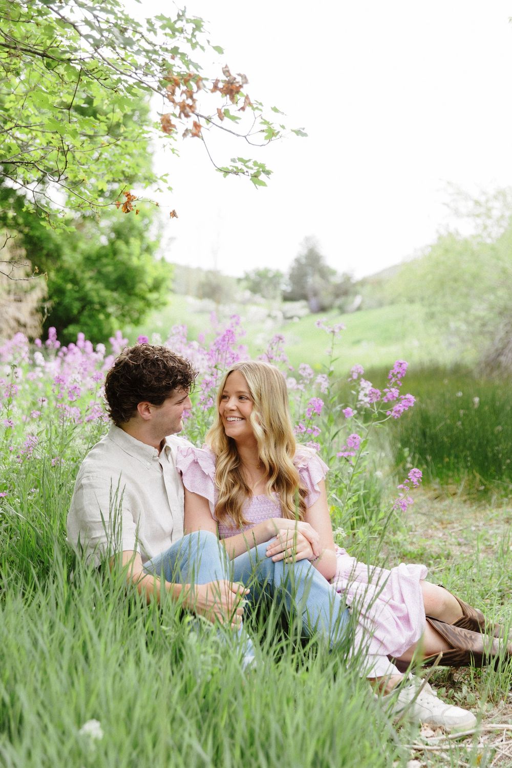 A couple sits together in a lush green meadow with purple wildflowers, smiling at each other on a warm spring day.