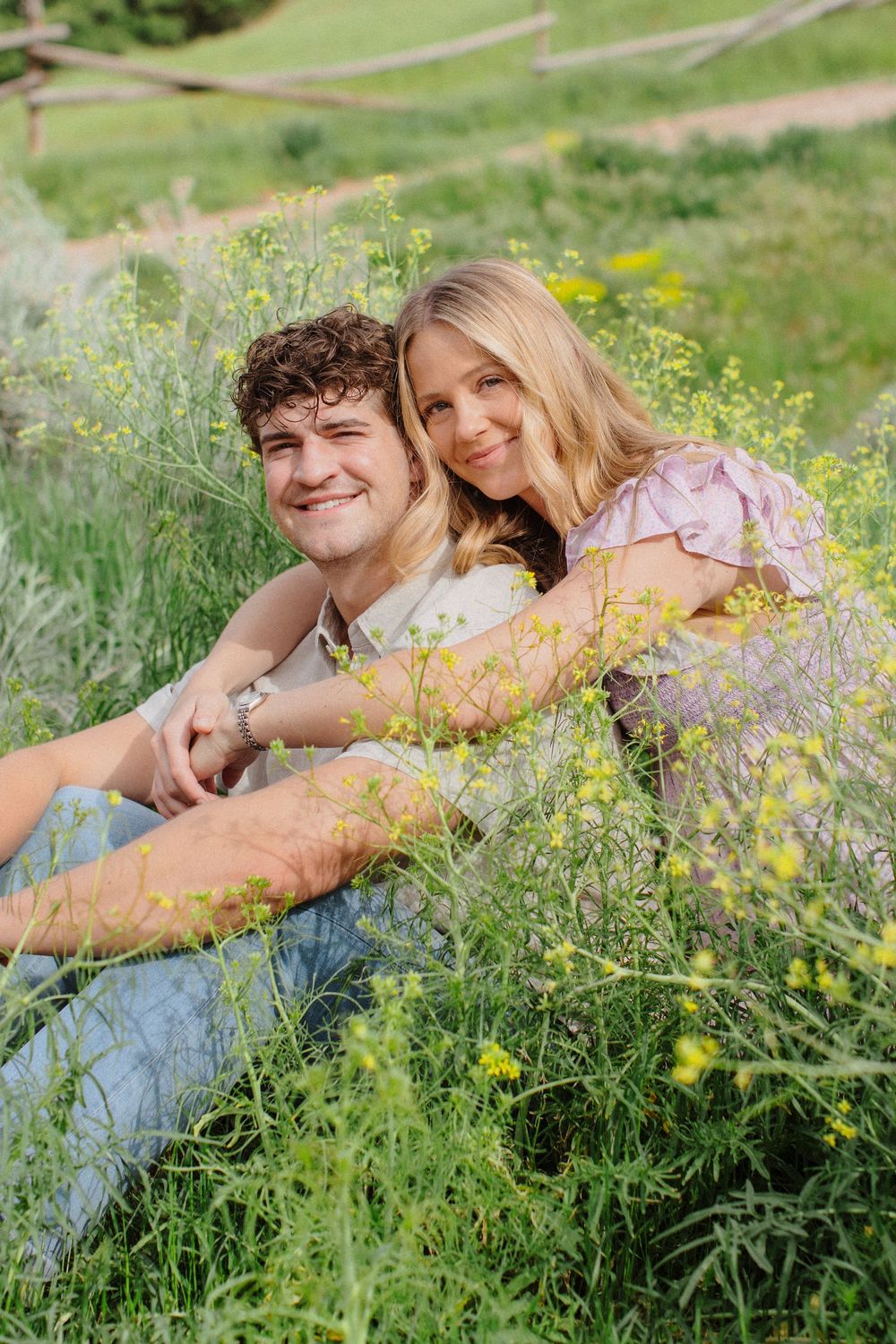 A young couple sits together in a field of yellow wildflowers, smiling warmly in a lush green outdoor setting.