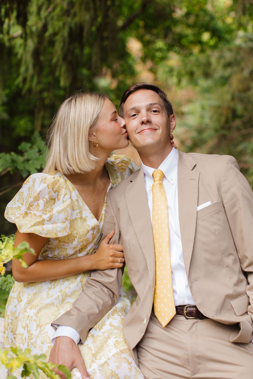 A woman in a floral dress kisses a smiling man in a tan suit with yellow tie outdoors.