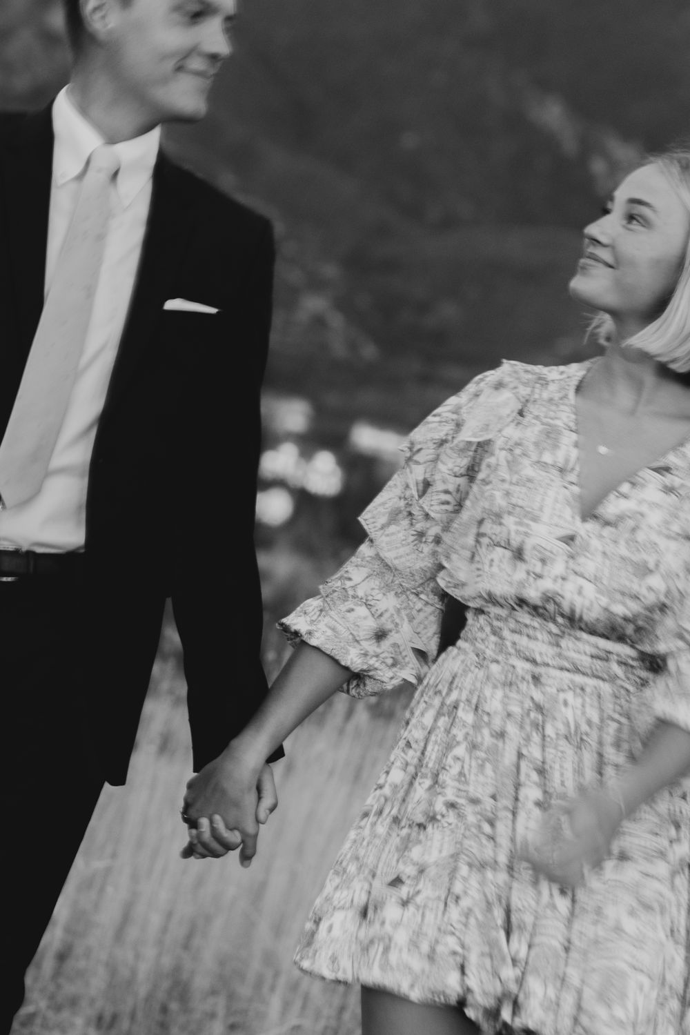 Black and white photo of a smiling couple holding hands, woman in floral dress gazing at man in suit.
