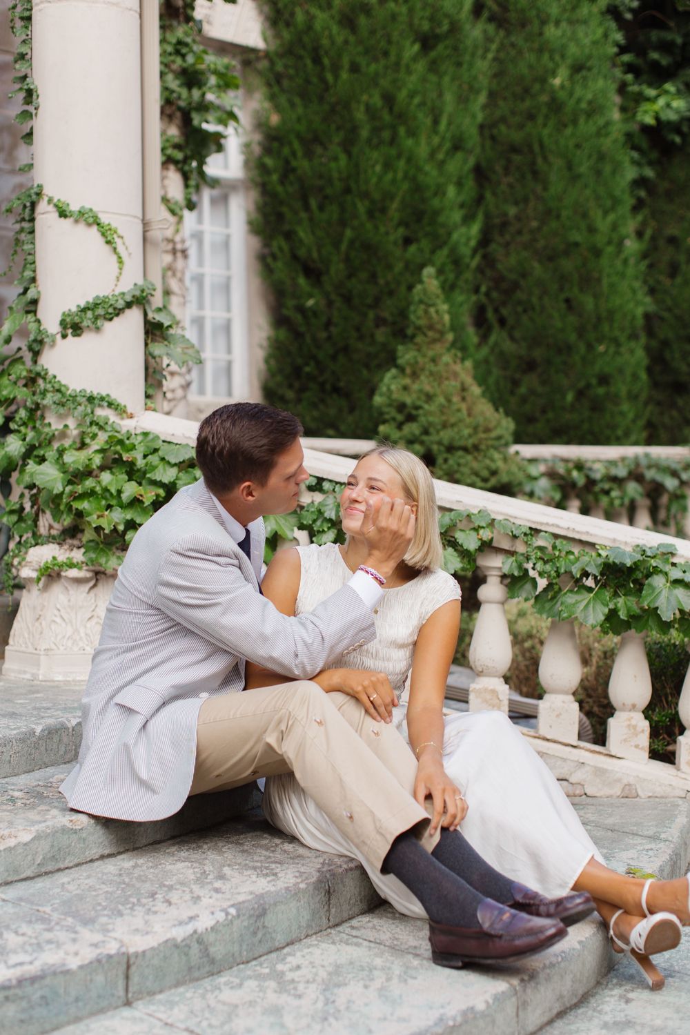 A couple sits on stone steps, smiling and touching faces, dressed in wedding attire with lush greenery behind them.