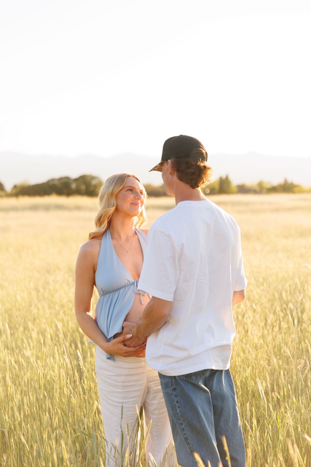 Pregnant couple holding baby bump in golden wheat field at sunset, woman in blue dress smiling at man in white tee.
