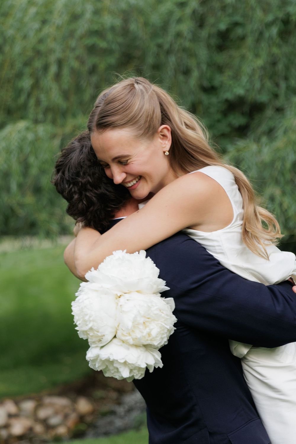 Bride smiling and hugging groom outdoors, holding white peony bouquet, with lush green weeping willow background.