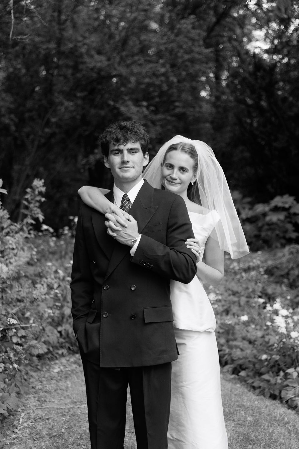 Black and white wedding portrait of bride and groom embracing on garden path, bride in veil hugging groom in suit.