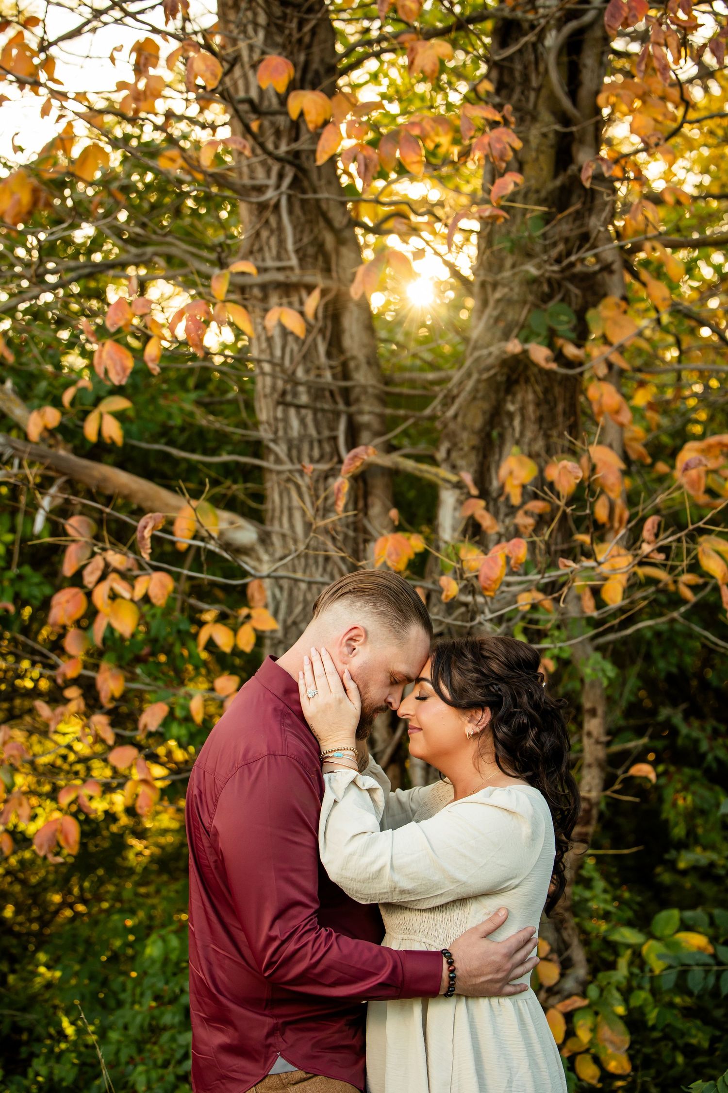 Couple embracing tenderly in autumn forest with golden foliage and warm sunset light filtering through colorful fall trees.