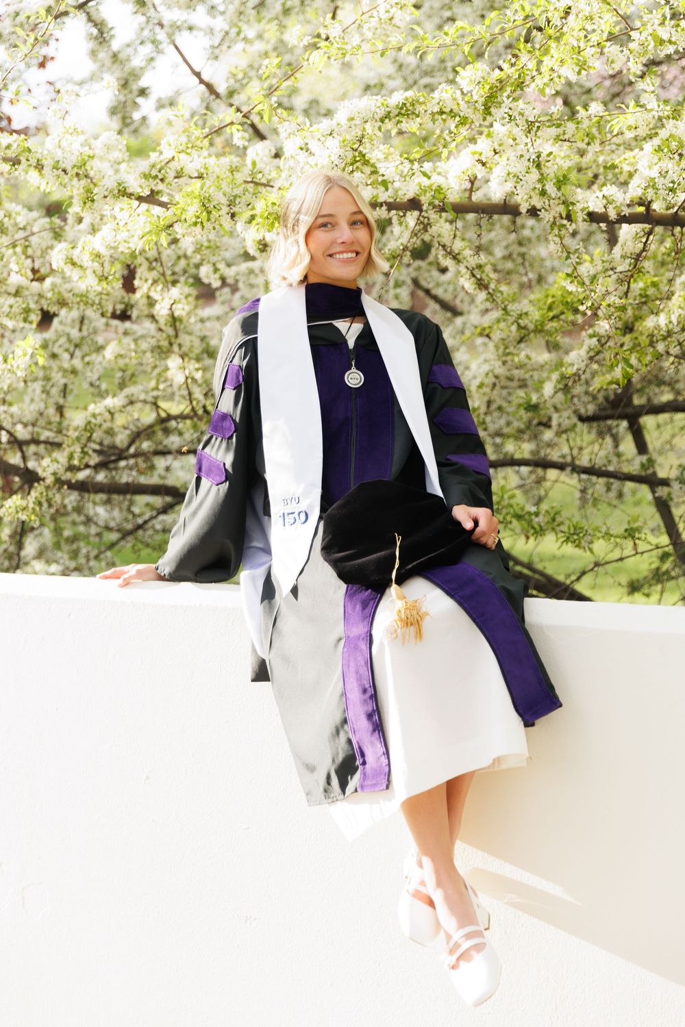 Blonde law school graduate in purple and black doctoral gown sitting on white wall among spring blossoms.