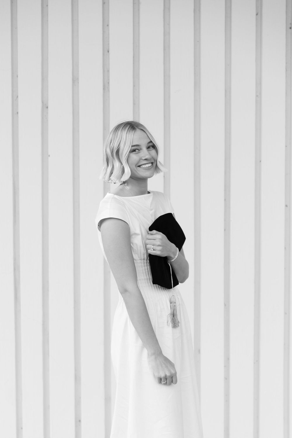 Black and white portrait of smiling blonde woman in white dress holding graduation cap outdoors.