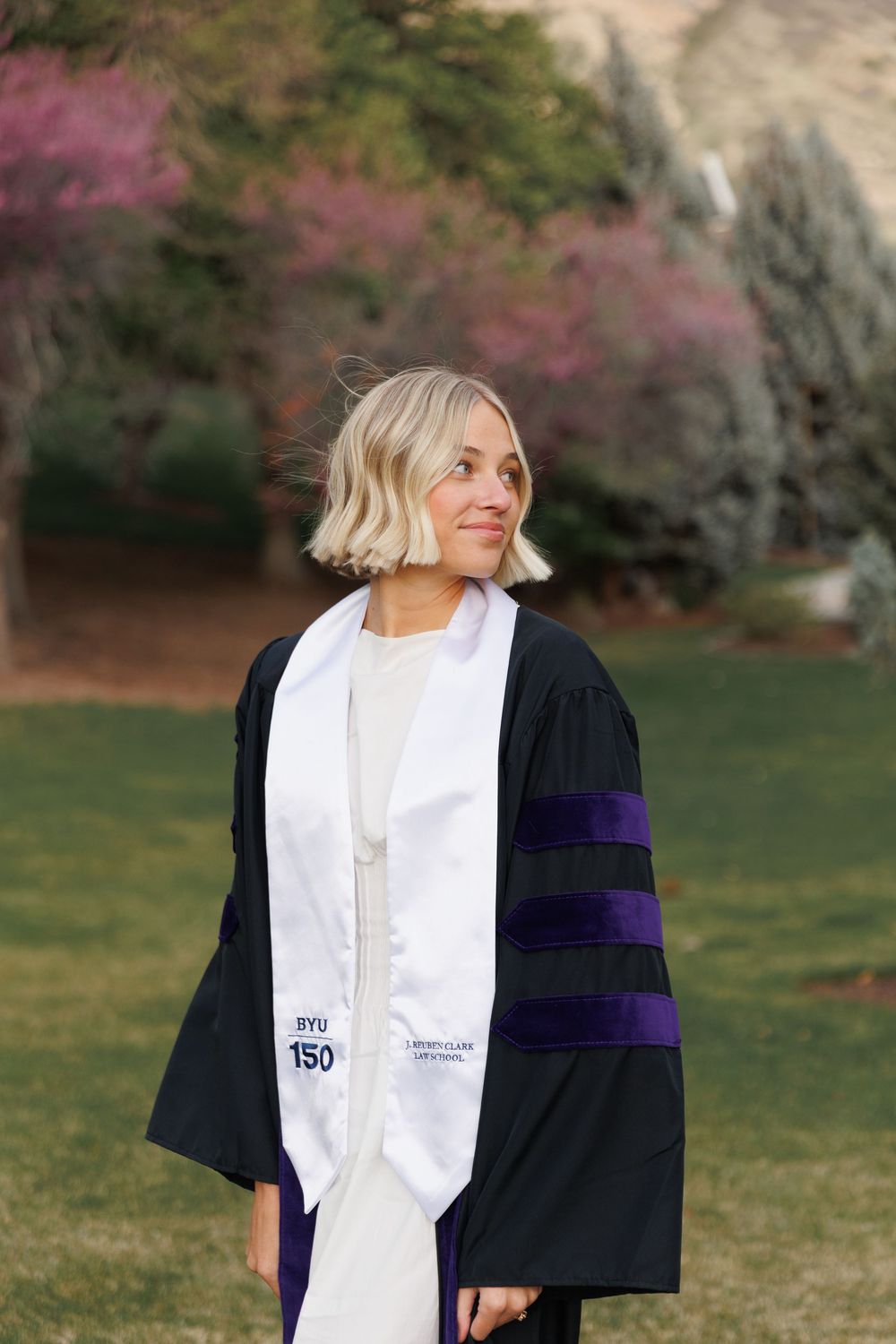 Blonde graduate wearing black doctoral gown with purple velvet stripes and BYU 150 white stole outdoors.