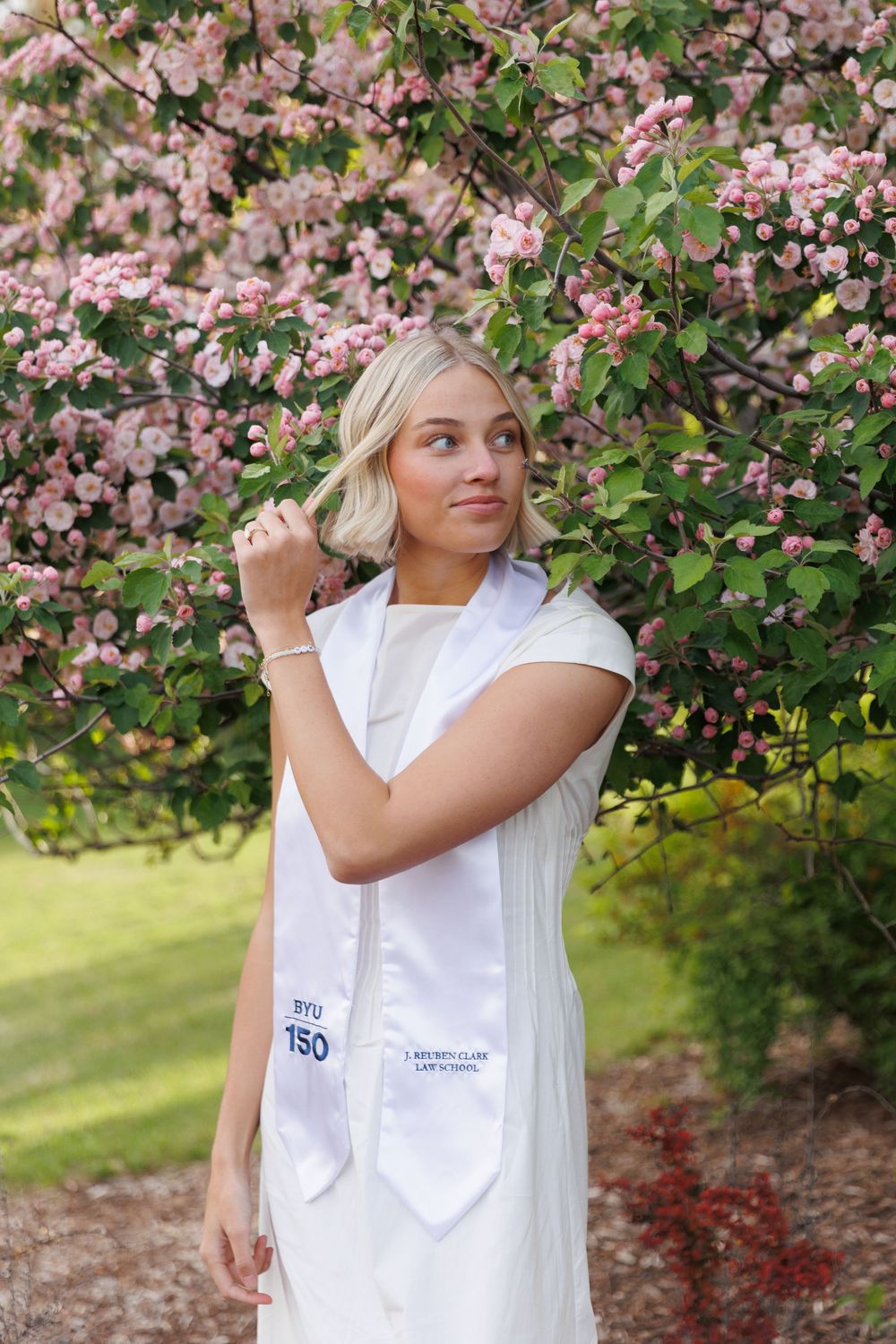 Blonde law school graduate in white dress and stole standing near pink flowering tree in garden.