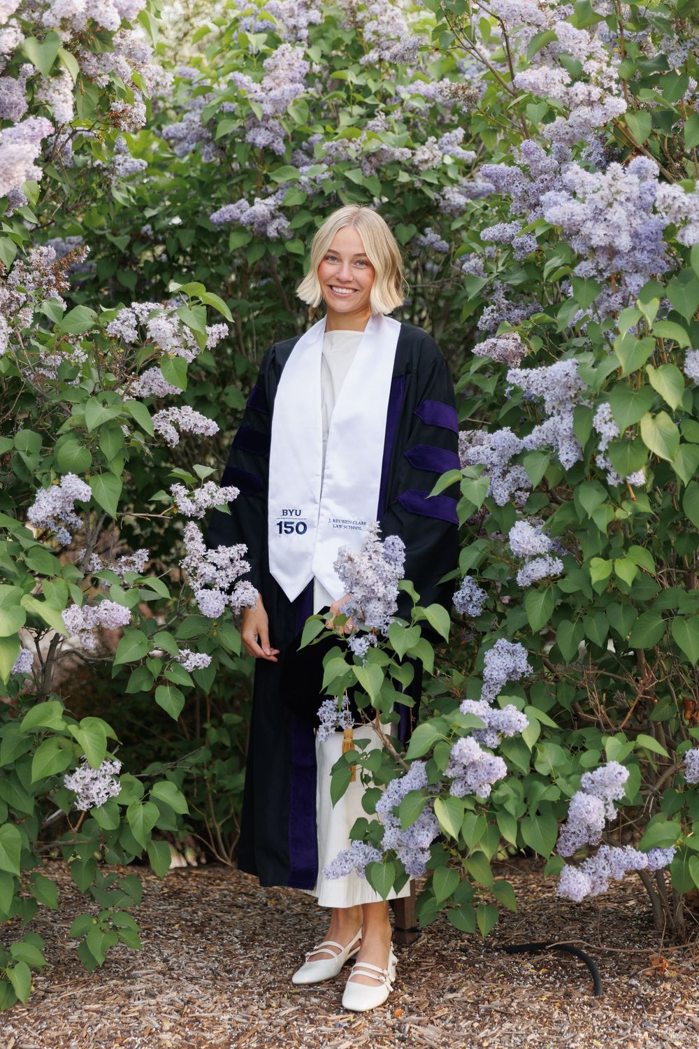 Smiling blonde graduate in navy doctoral gown and white BYU stole standing among purple lilac bushes.