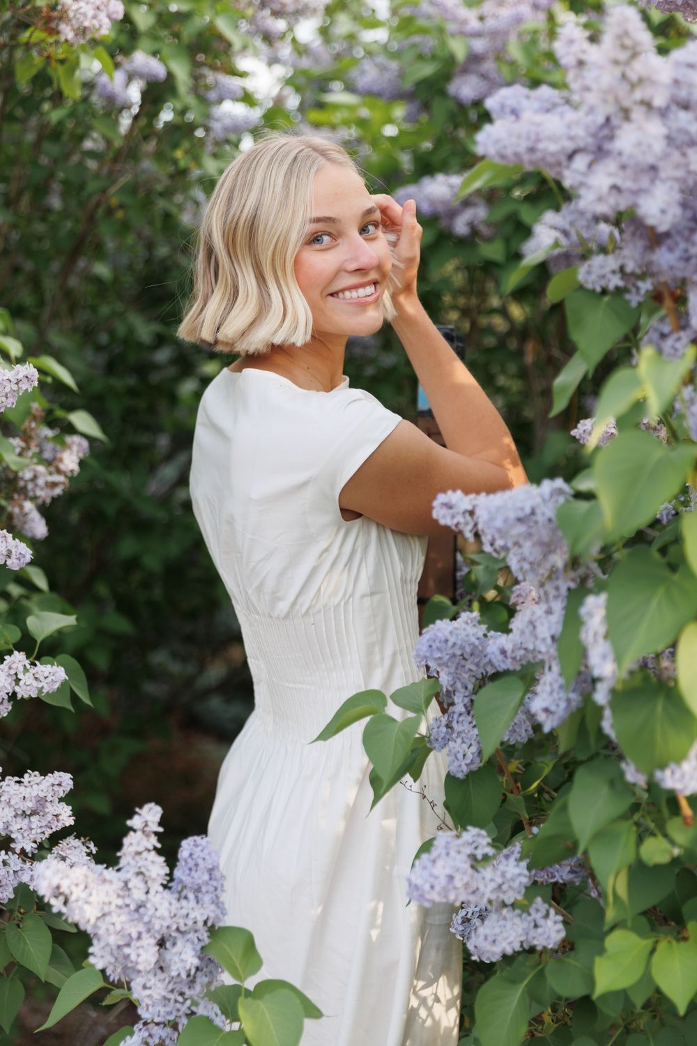 Smiling blonde woman in white dress surrounded by blooming purple lilac flowers in a garden.