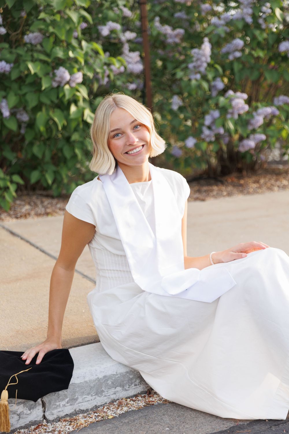Smiling blonde law school graduate in white dress and stole sitting on curb near lilac bushes.