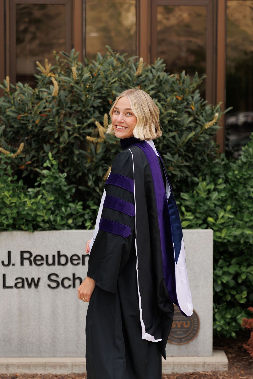 Smiling woman in law graduation gown posing outside BYU J. Reuben Clark Law School sign.