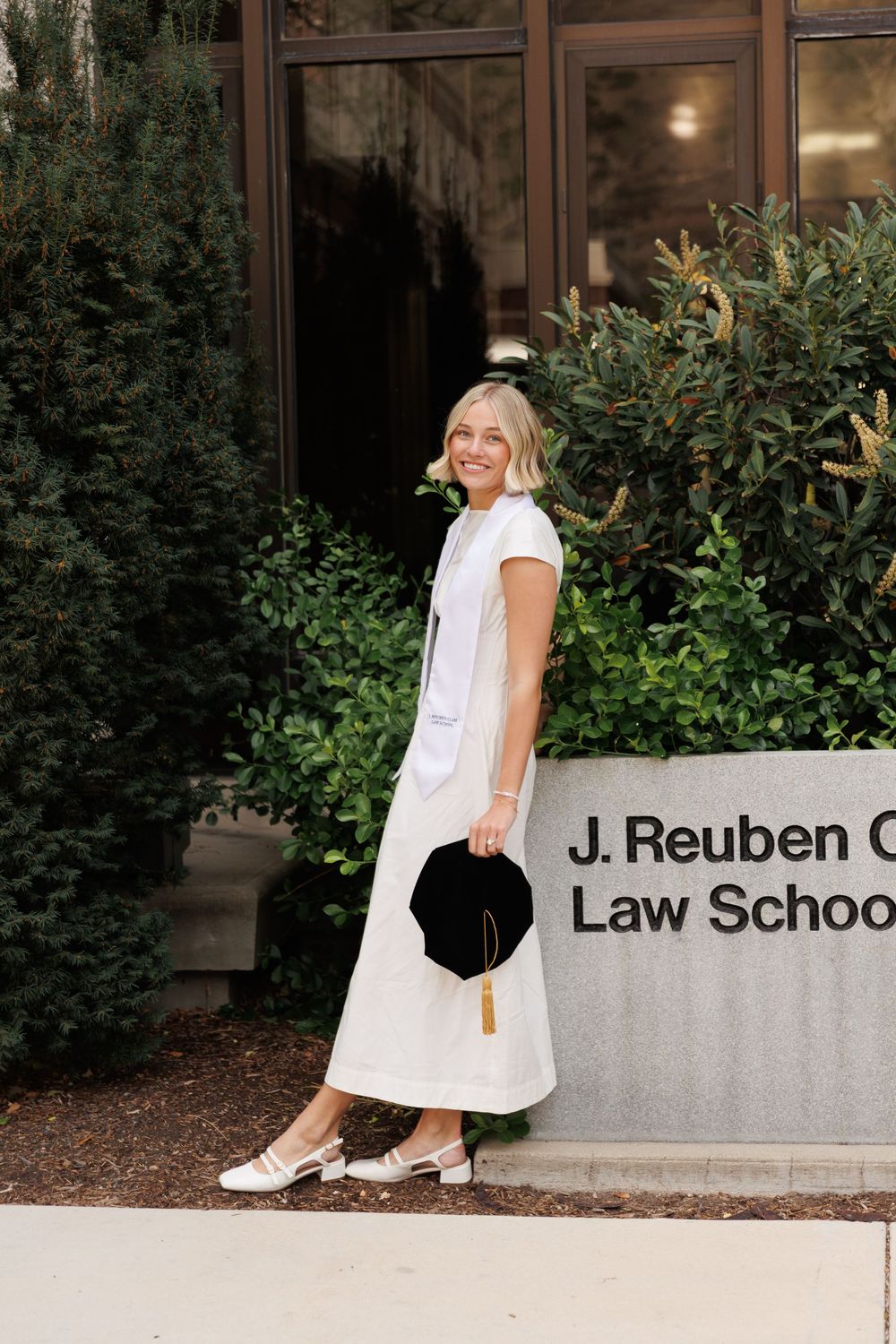 Smiling blonde law school graduate in white dress holding black cap outside J. Reuben Clark Law School.