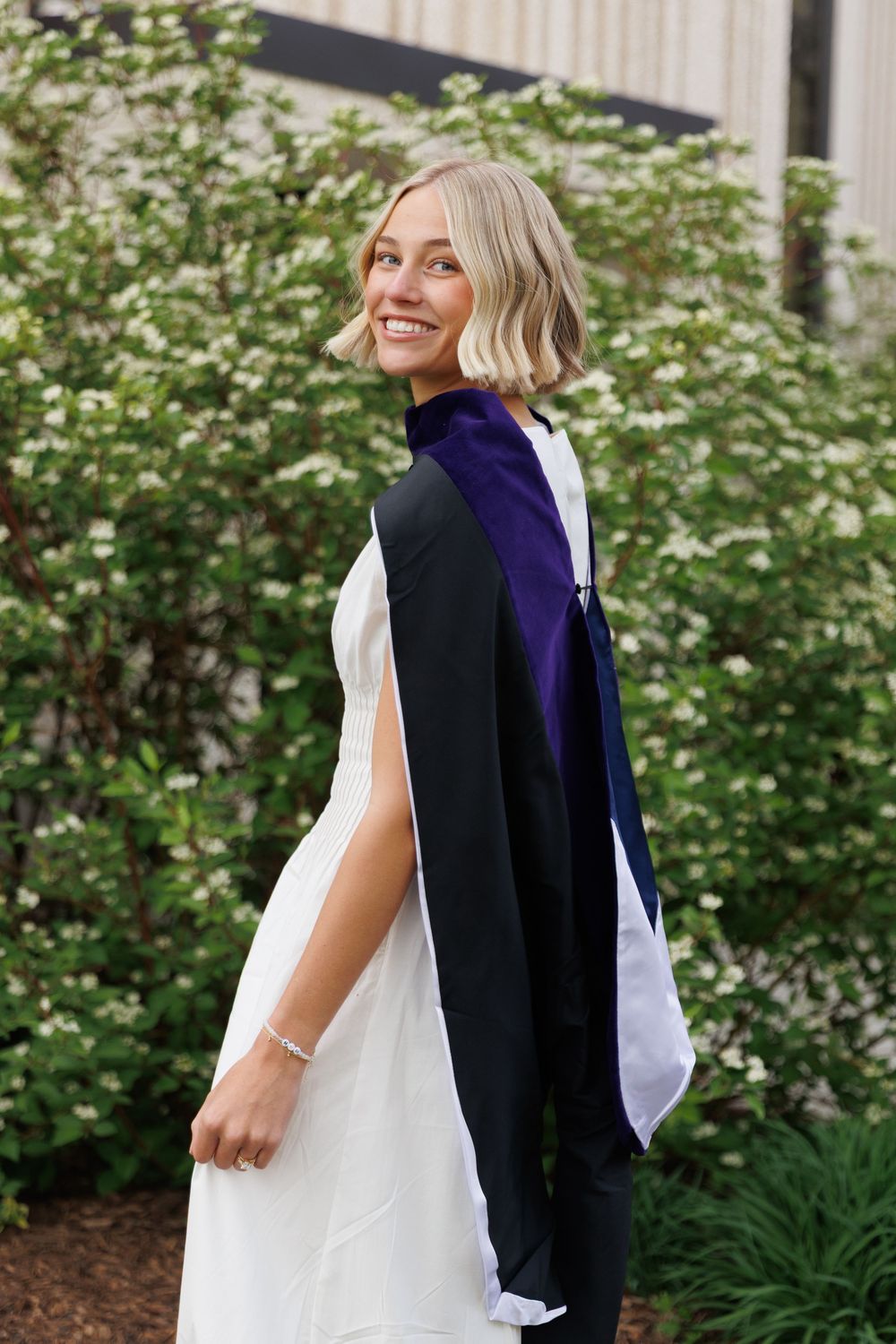 Blonde woman in black graduation gown with purple hood smiling outdoors among white flowering bushes.
