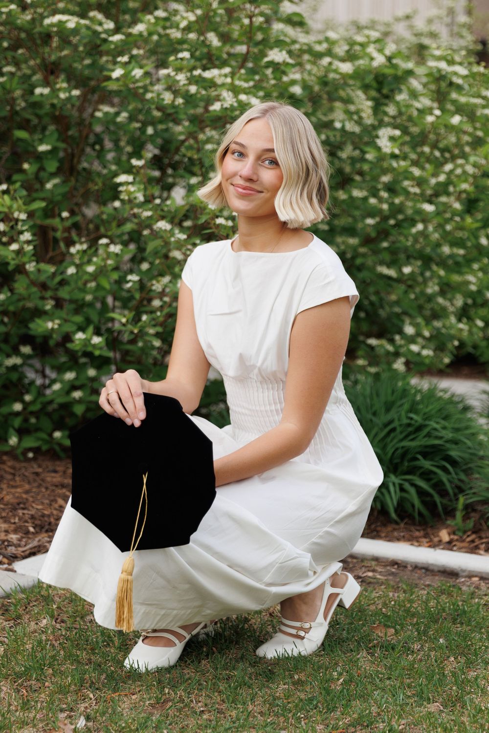Blonde graduate in white dress crouching on grass holding black mortarboard cap with gold tassel.