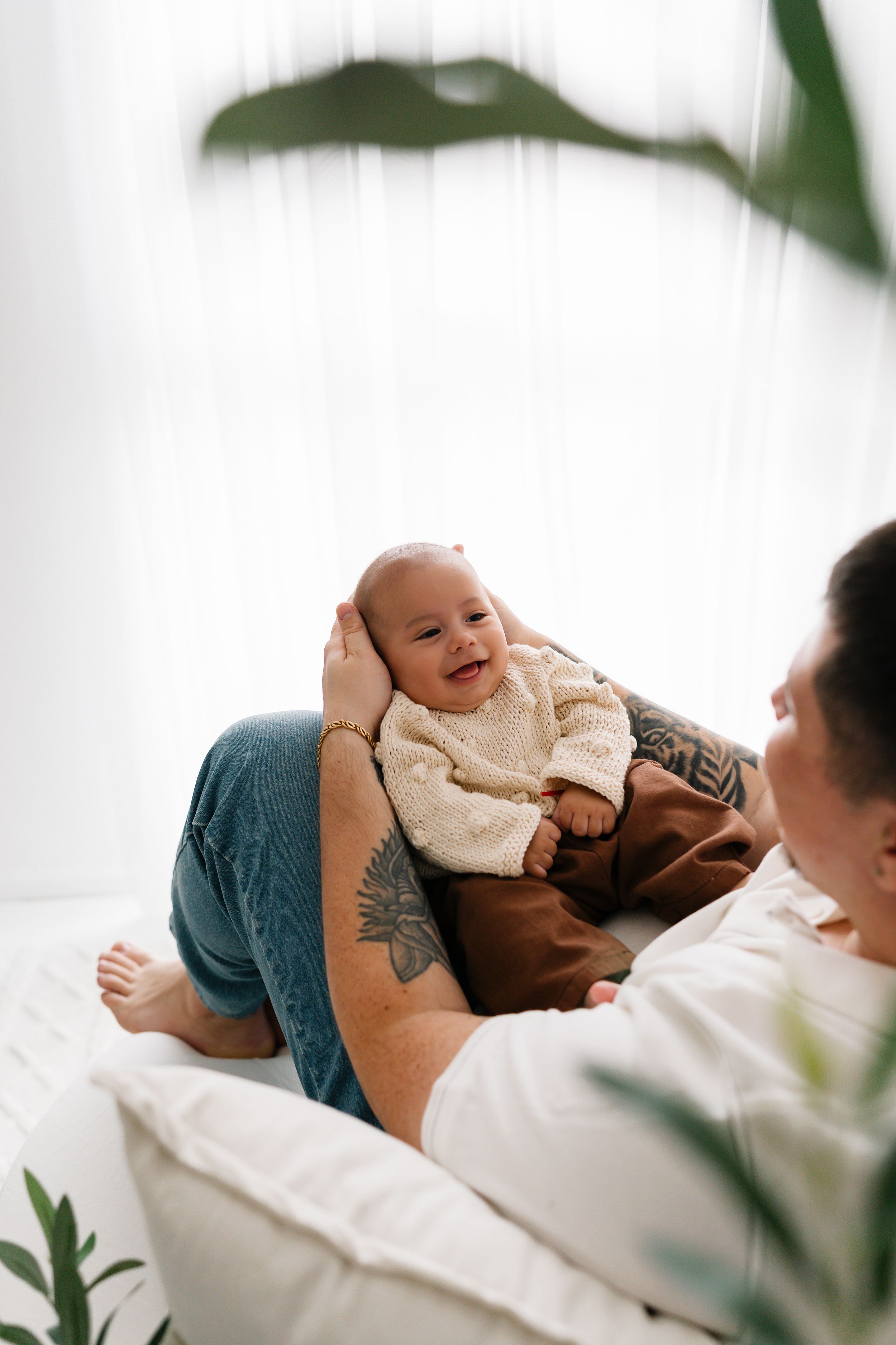 Tattooed parent lying on bed holding smiling baby in cream sweater, with green plants and white curtains in background.