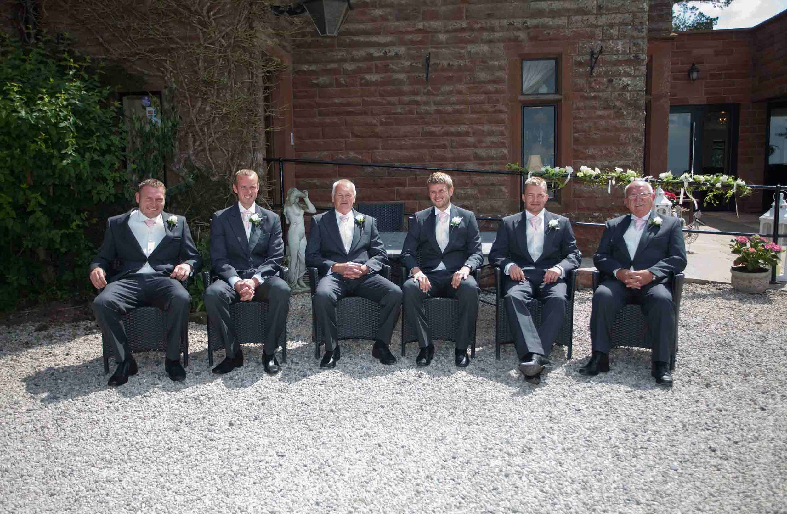 Groom and wedding party in dark suits seated outside Heather Glen Country House Hotel, with the red brick building behind them.