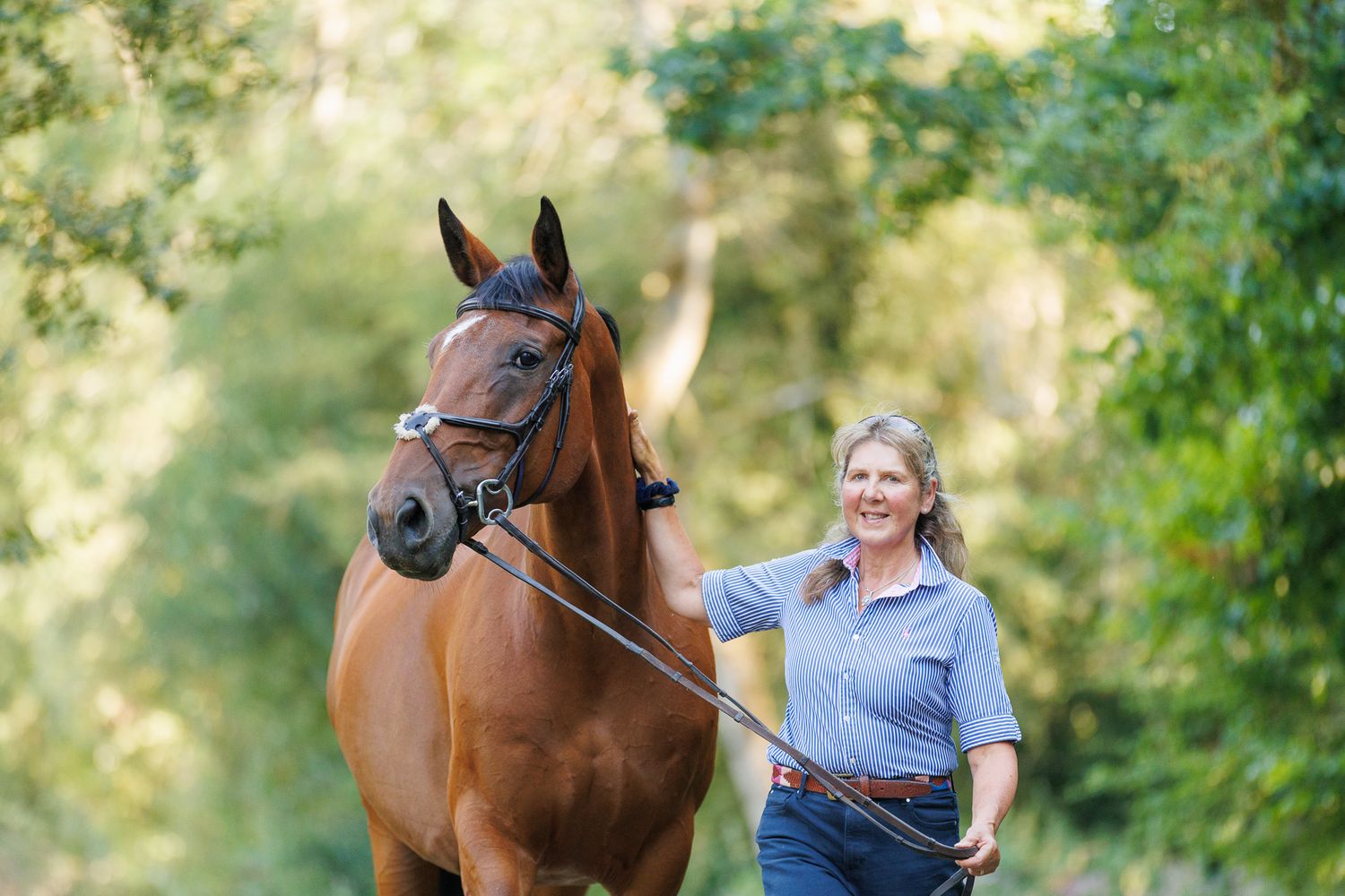 Woman in light blue gown walking with a bay horse on a sunlit tree-lined gravel path.
