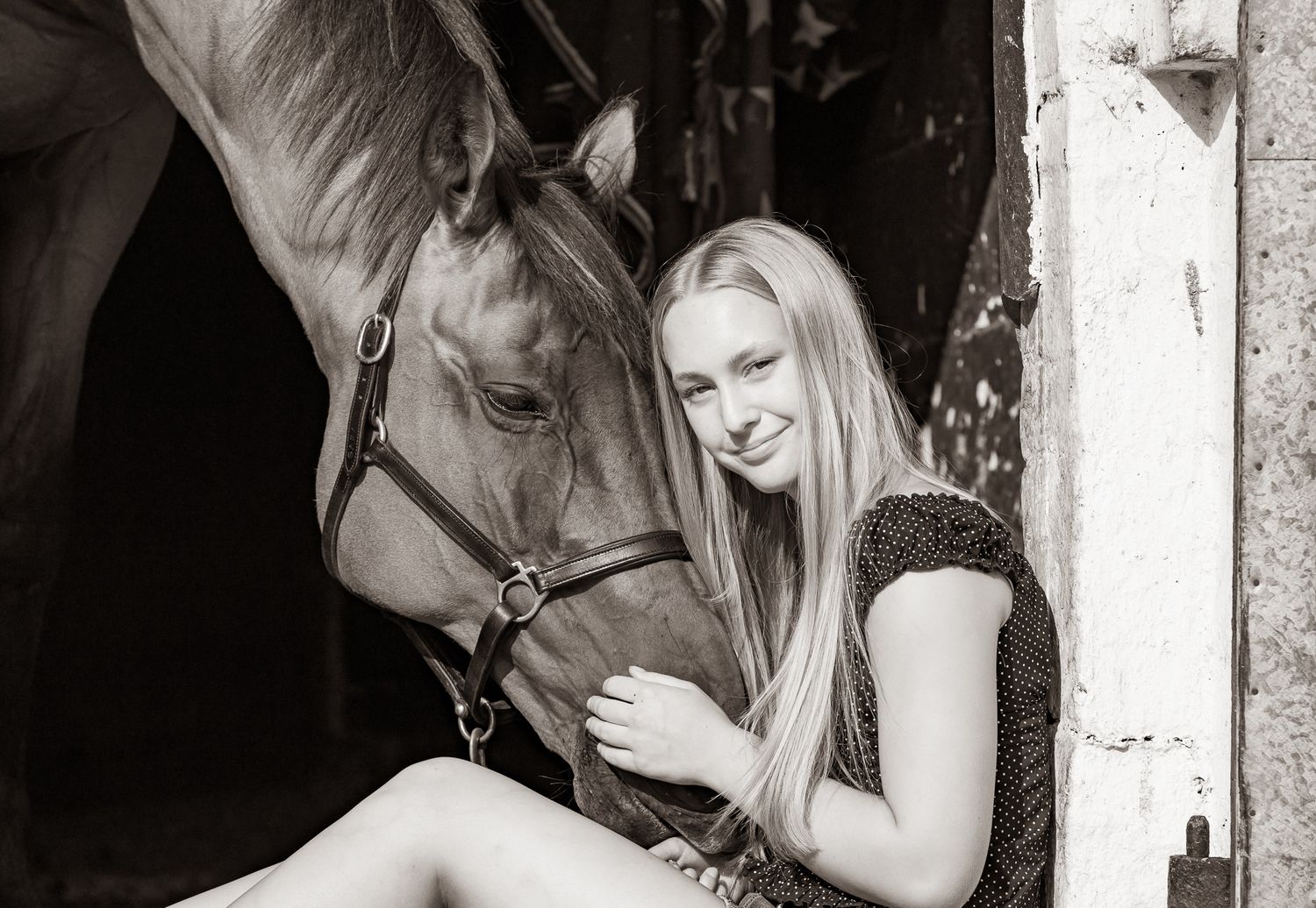 Young woman in black top smiling while gently embracing a brown horse outdoors among lush green trees.